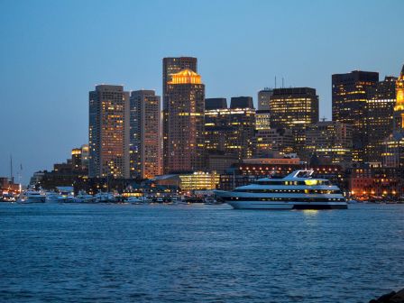 A cityscape at dusk with illuminated buildings in the background and a large boat sailing on a body of water in the foreground.