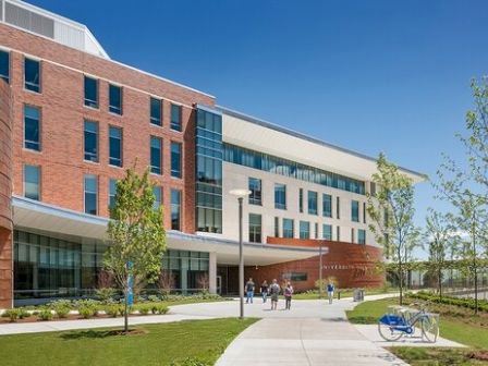 The image shows a modern multi-story building with large windows, surrounded by greenery. A few people walk on the path towards the entrance.