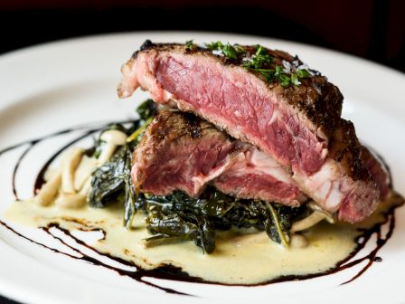 A plate of rare steak served on a bed of greens with a creamy sauce and balsamic reduction garnish.