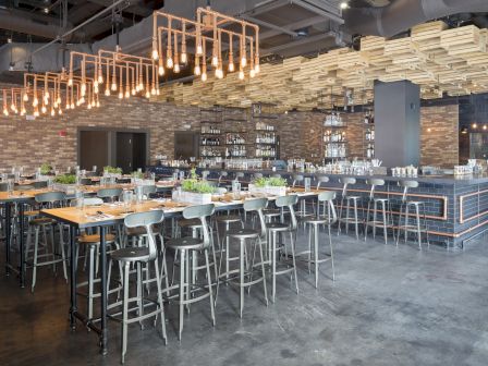 A modern restaurant interior with stylish lighting, high tables and chairs, and an elaborate bar area showcasing bottles.