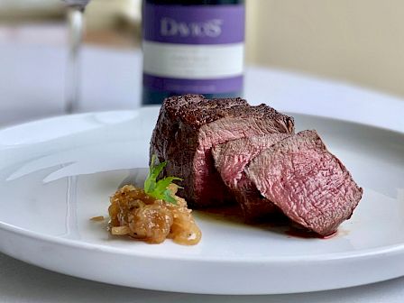 A plate with sliced steak and a small garnish, accompanied by a glass of red wine and a wine bottle in the background.