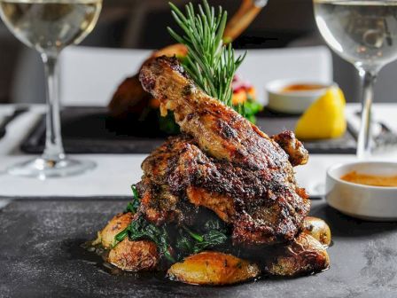 A beautifully plated dish of roasted chicken with herbs, potatoes, and greens, accompanied by two glasses of white wine and dipping sauce.