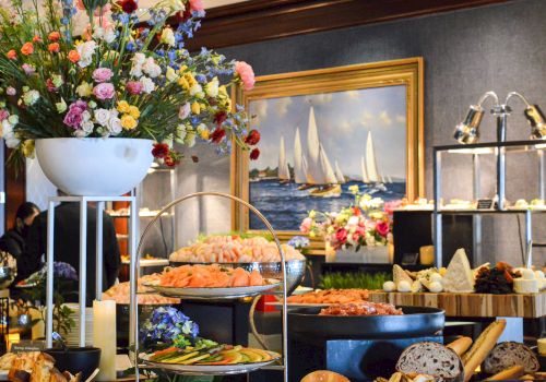 A buffet table is adorned with various foods, including seafood and bread, alongside elegant flower arrangements and a painting of sailboats in the background.