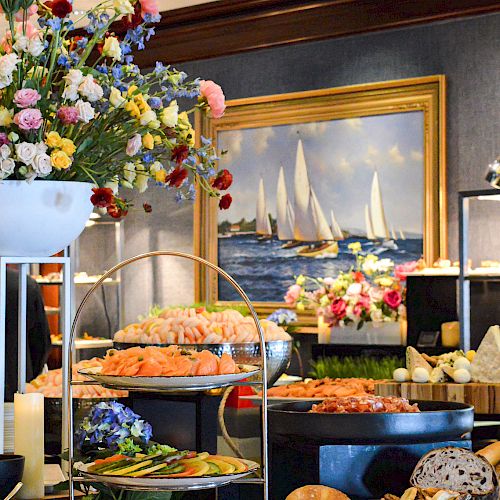 A buffet table is adorned with various foods, including seafood and bread, alongside elegant flower arrangements and a painting of sailboats in the background.