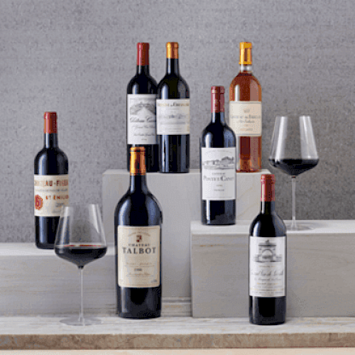 The image displays seven wine bottles of various labels, with two glasses partially filled with red wine on a stepped gray platform.