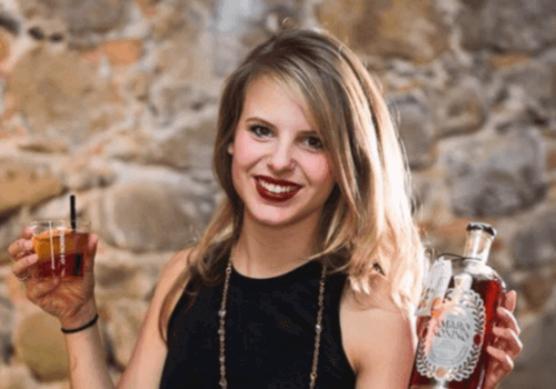 A woman with blonde hair is holding a glass with a drink in one hand and a bottle of liquor in the other, smiling in front of a stone wall.