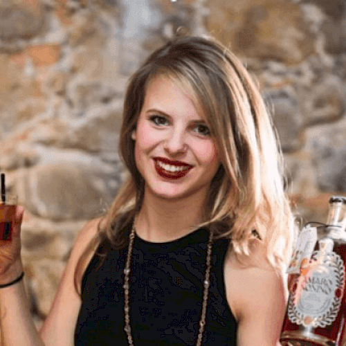 A woman with blonde hair is holding a glass with a drink in one hand and a bottle of liquor in the other, smiling in front of a stone wall.