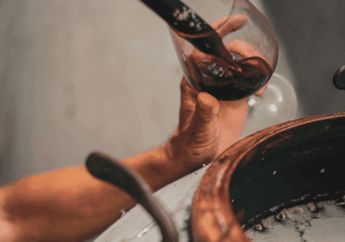 A hand holding a wine glass receiving wine from a vessel, possibly in a wine-making setting.