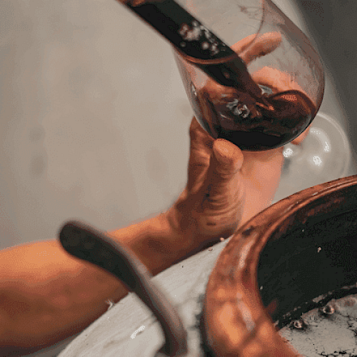 A hand holding a wine glass receiving wine from a vessel, possibly in a wine-making setting.