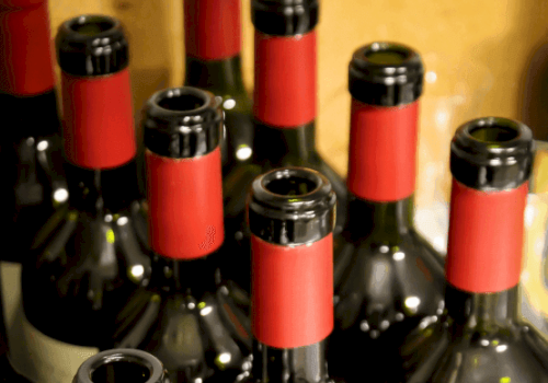 The image shows multiple empty wine bottles with red and black neck bands, placed closely together, likely in a storage area.