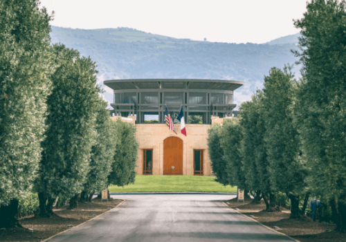 The image shows a modern building surrounded by trees, with a road leading to it. American and French flags are displayed in front of the building.