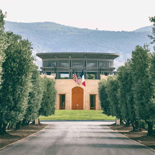 The image shows a modern building surrounded by trees, with a road leading to it. American and French flags are displayed in front of the building.