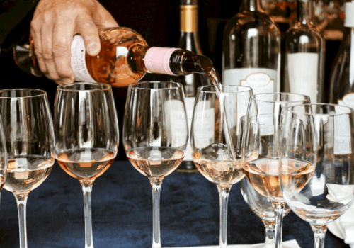 A person is pouring rosé wine into several wine glasses, with various wine bottles in the background.