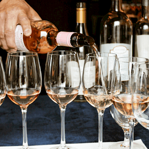 A person is pouring rosé wine into several wine glasses, with various wine bottles in the background.