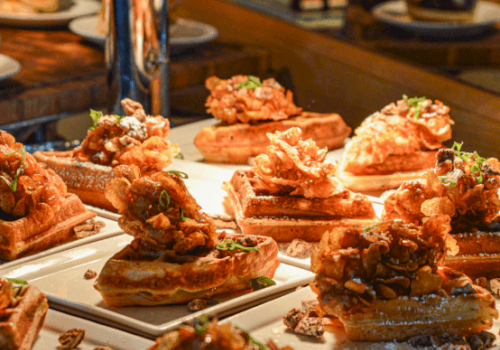 A buffet display of chicken and waffles garnished with herbs, arranged on plates under warm lighting, ready to be served.