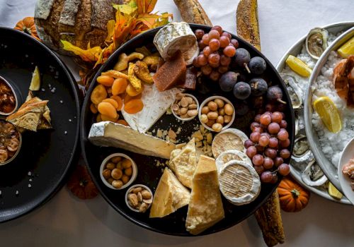 An assortment of cheeses, grapes, nuts, dried fruits, and bread are displayed on a platter alongside dishes of seafood and lemon wedges.