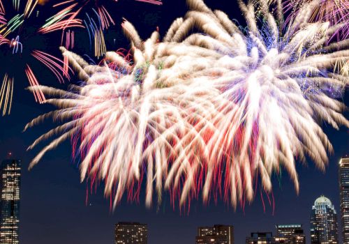 A city skyline at night with a vibrant fireworks display illuminating the sky and reflecting on a body of water.