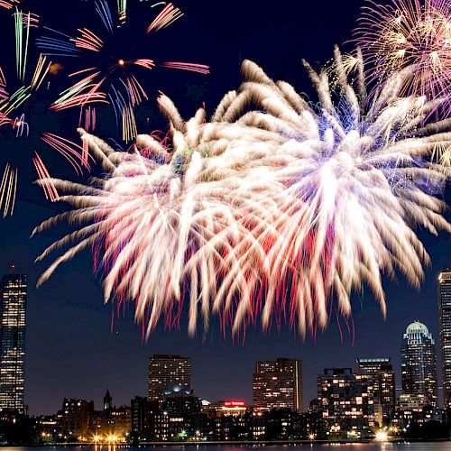 A city skyline at night with a vibrant fireworks display illuminating the sky and reflecting on a body of water.