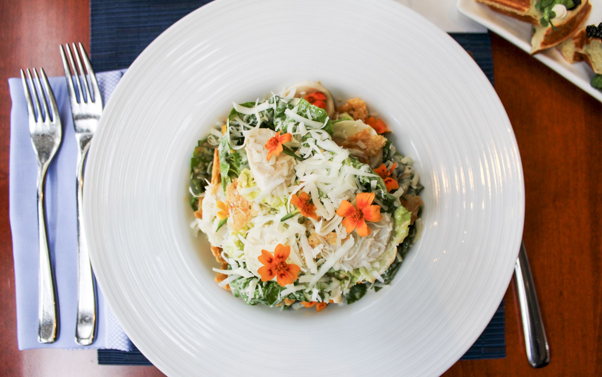 A plate of salad topped with shredded cheese and edible flowers, with a fork and knife placed on a napkin beside it.