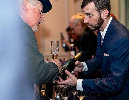 A man in a suit is pouring a beverage into a glass held by an older man wearing a hat, during what appears to be a social gathering.