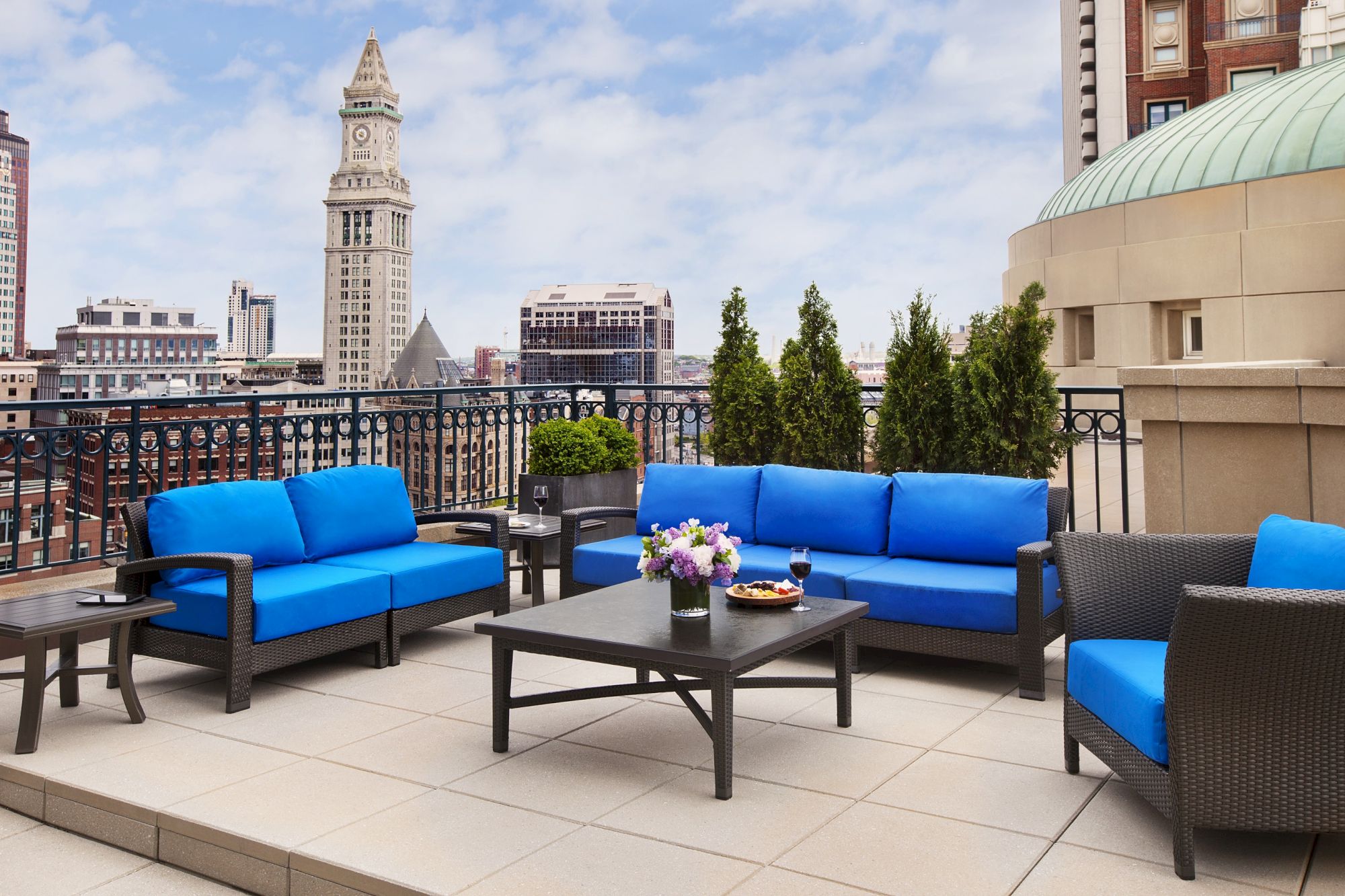 A rooftop patio with blue cushioned seating, a black coffee table with food, and a cityscape view featuring a clock tower in the background.