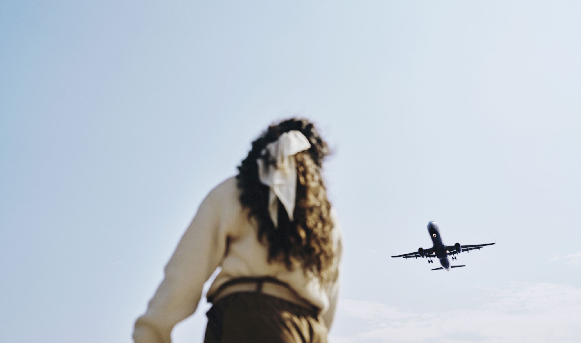 An individual with long hair and a white ribbon looks up as an airplane flies overhead against a clear sky.