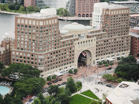 An aerial view of a large, archway-adorned building near a waterway, surrounded by greenery, pathways, and neighboring structures, with people visible.