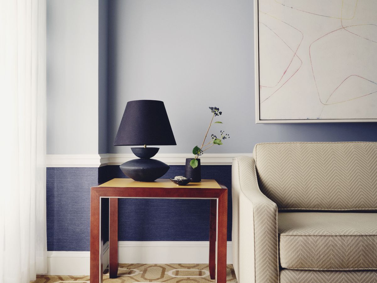 The image shows a living room corner with a beige couch, a wooden side table holding a black lamp, and a plant. Modern art decorates the wall above.