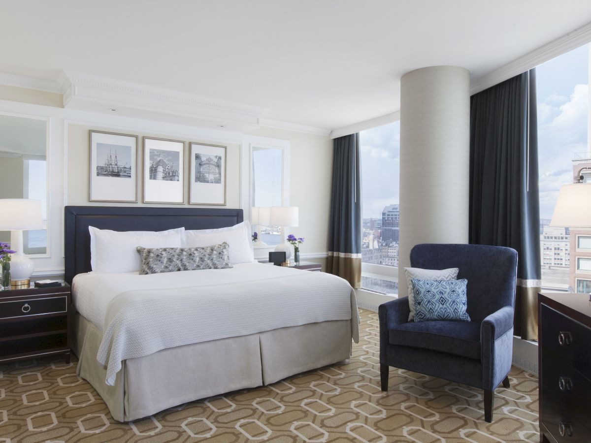 A modern, well-lit hotel room with a large bed, bedside tables with lamps, a blue armchair, and large windows showcasing a city view.