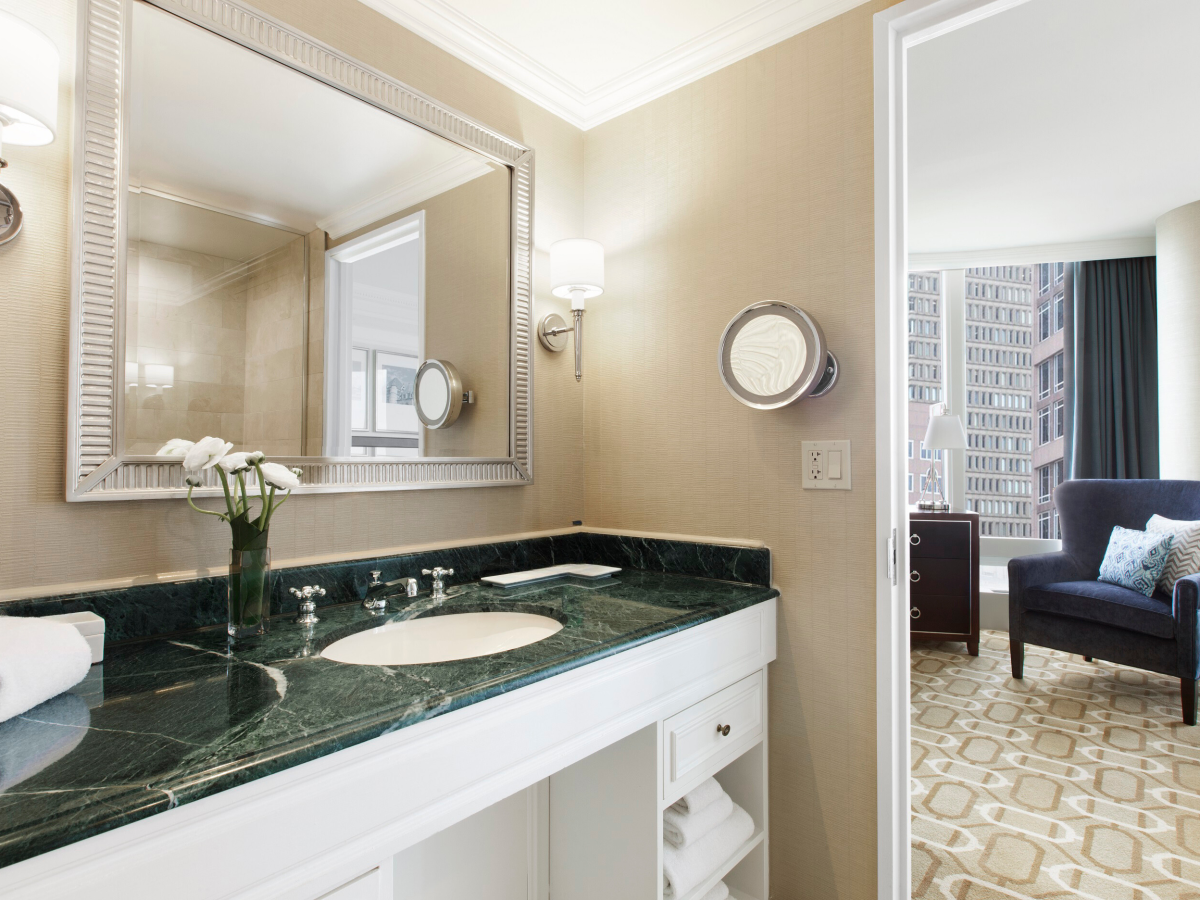 A bathroom with a green marble countertop, sink, mirror, and vanity lights, opening into a room with a chair and city view.