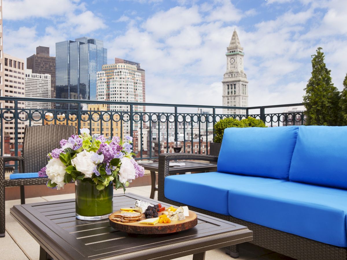 A rooftop patio with a blue sofa, a coffee table with flowers and a food tray, overlooking a city skyline with tall buildings.