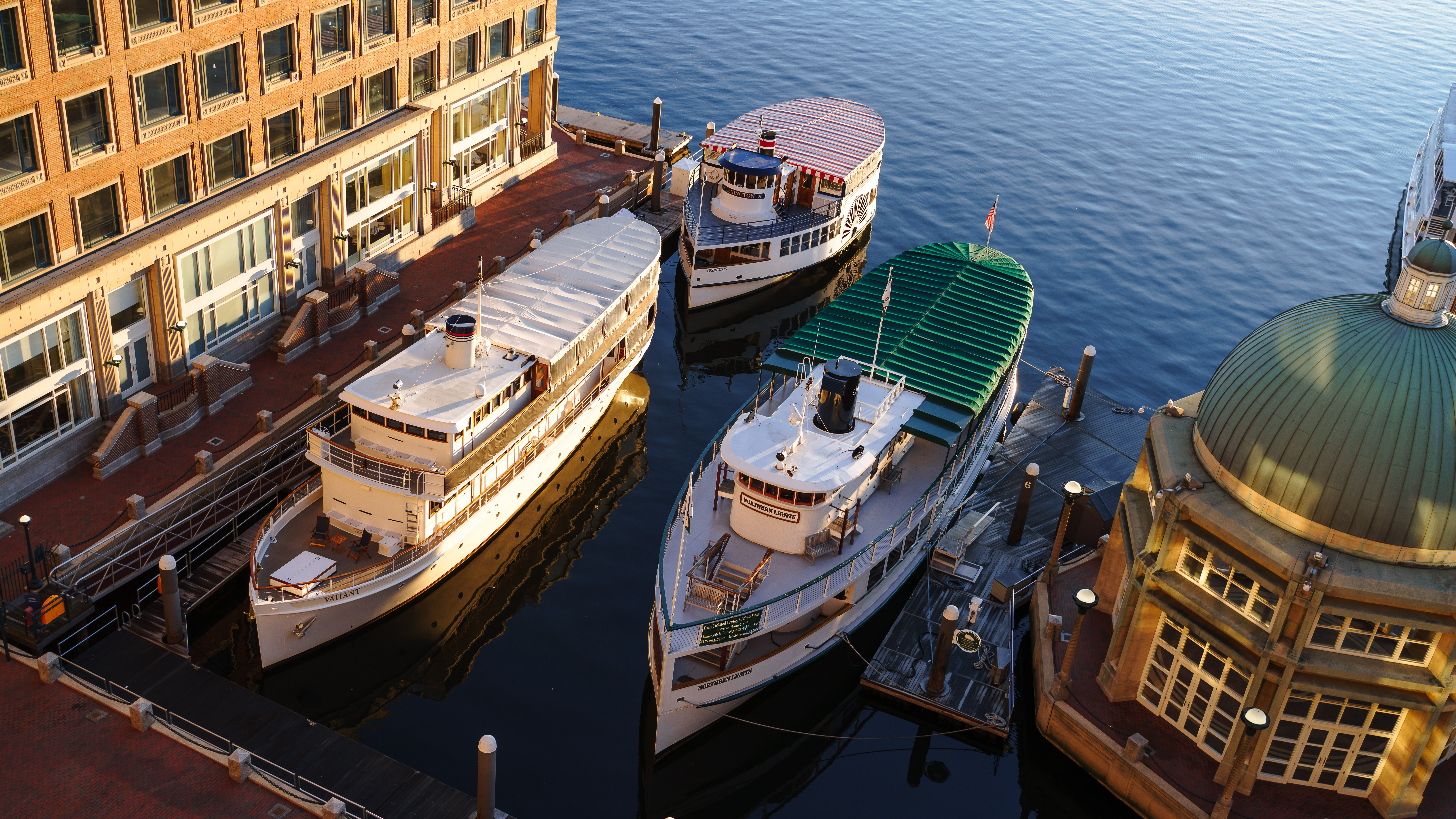 Three boats are docked by a waterfront building. The boats are aligned parallel to each other. The scene takes place at sunset.