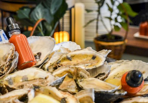 An assortment of oysters on a platter, adorned with lemon slices, and accompanied by two dropper bottles containing a red liquid.