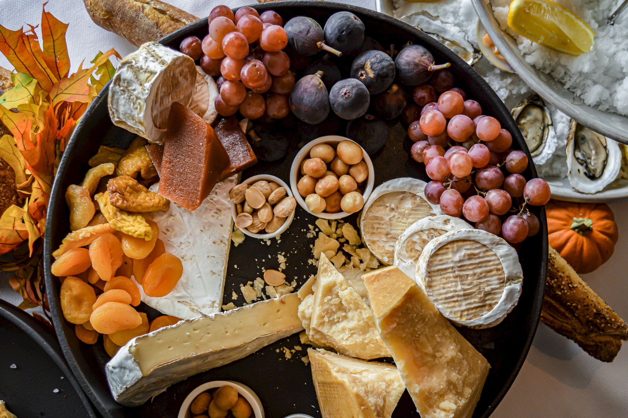 A cheese platter with various cheeses, grapes, nuts, and dried apricots, accompanied by bread and oysters on the side.