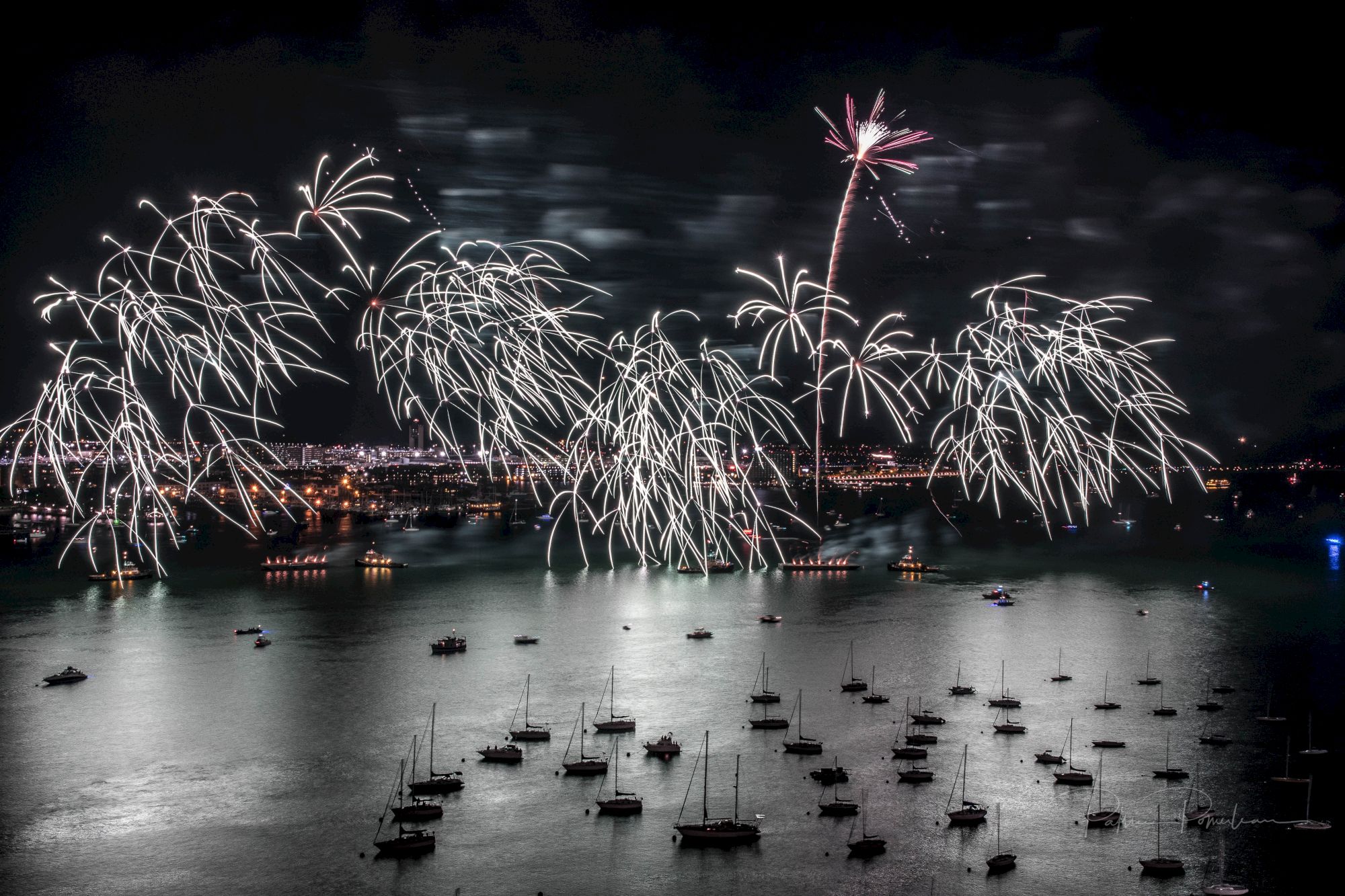 A night scene with fireworks lighting up the sky over a body of water filled with boats.