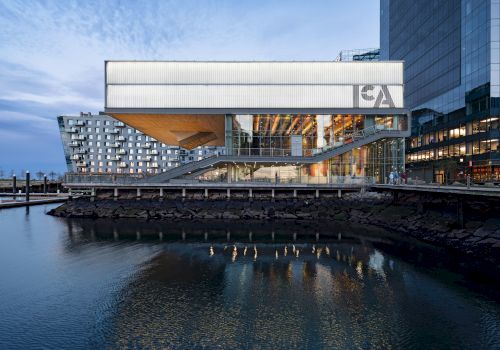 A modern, multi-story building with large glass windows is situated by a waterfront, featuring the letters 