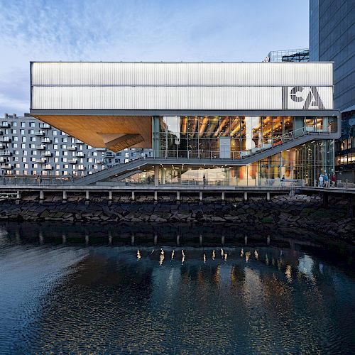 A modern, multi-story building with large glass windows is situated by a waterfront, featuring the letters 