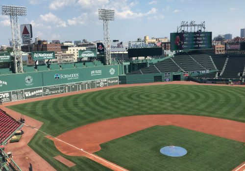 The image shows an empty baseball stadium with green seats, the field, and a batting cage set up, under a partly cloudy sky, with the city in the background.