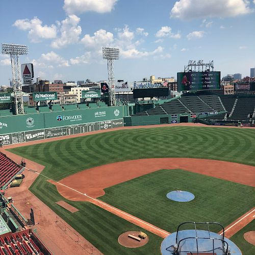 The image shows an empty baseball stadium with green seats, the field, and a batting cage set up, under a partly cloudy sky, with the city in the background.