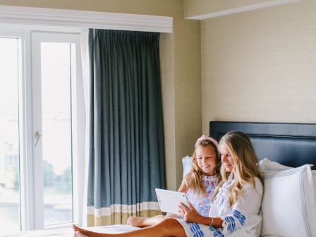 A woman and a child are sitting on a bed in a hotel room, looking at a tablet. The room has a large window with curtains.