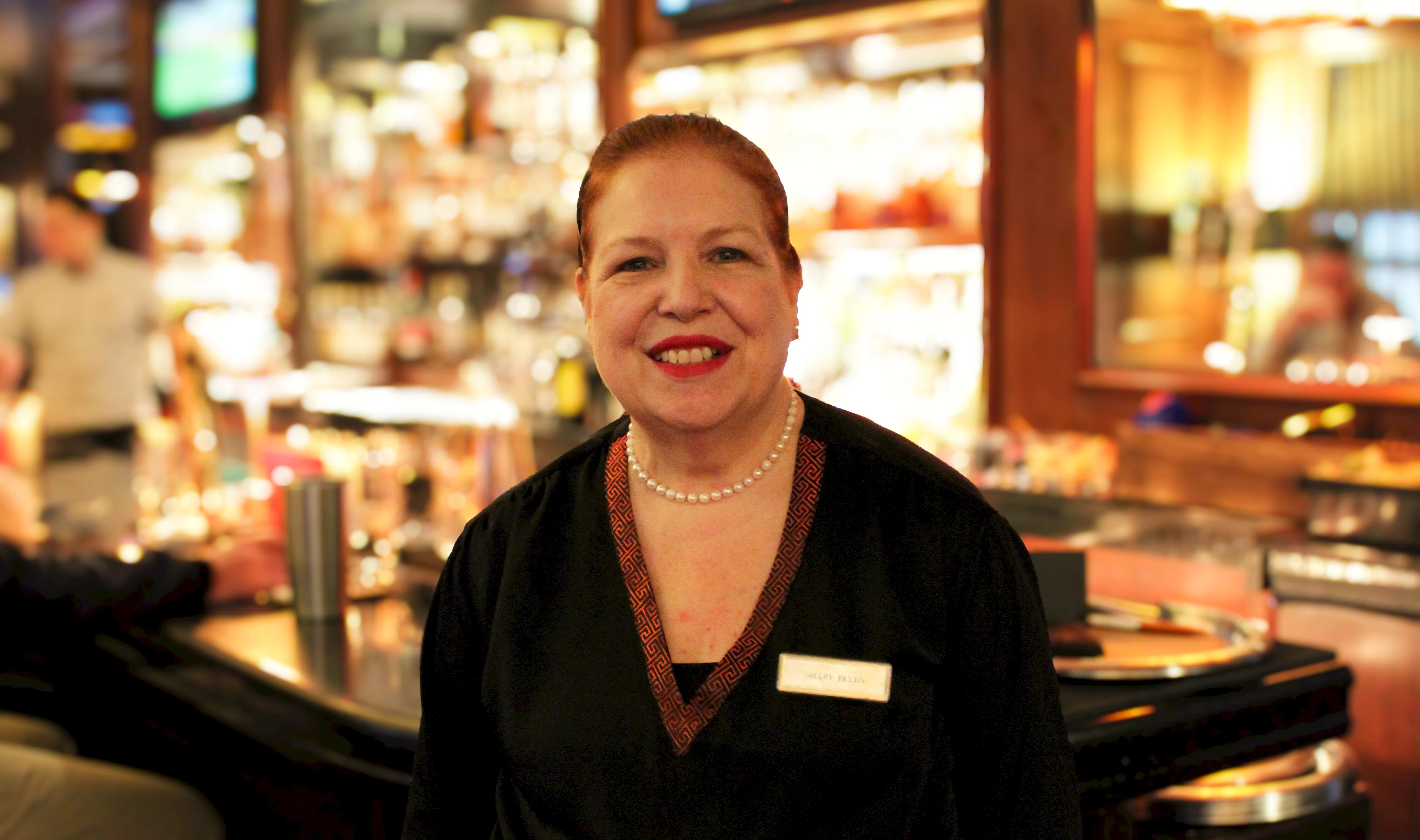 A smiling person stands in a warm, dimly lit bar or restaurant, wearing a name tag and dark clothing, with a fully stocked bar in the background.