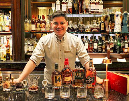 A bartender stands behind a bar filled with various bottles of alcohol, glasses, and a cocktail shaker, smiling.