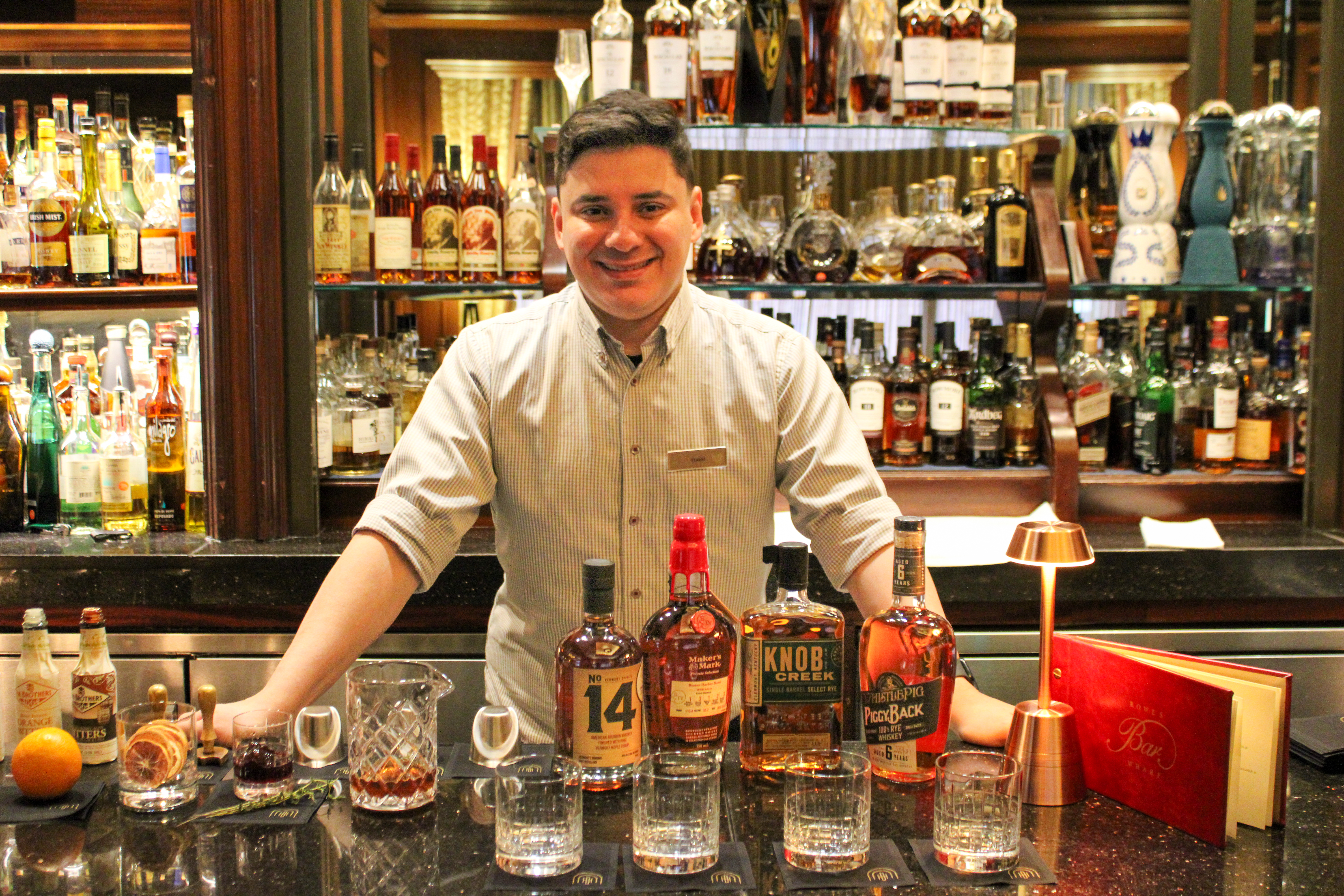 A bartender stands behind a bar filled with various bottles of alcohol, glasses, and a cocktail shaker, smiling.