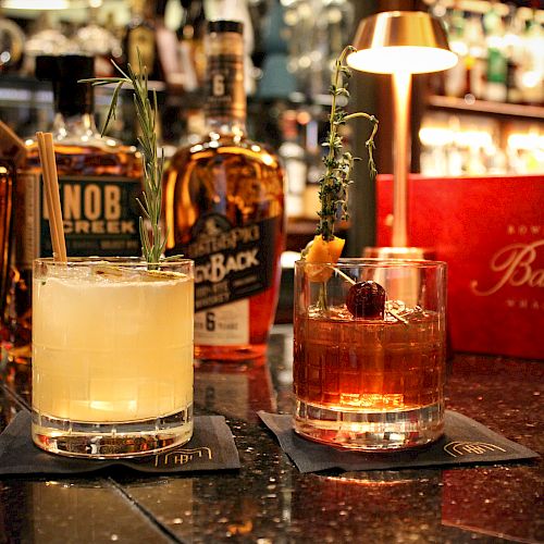 Two cocktails garnished with herbs on a bar counter, surrounded by bottles of bourbon and whiskey, with a dimly lit ambiance.