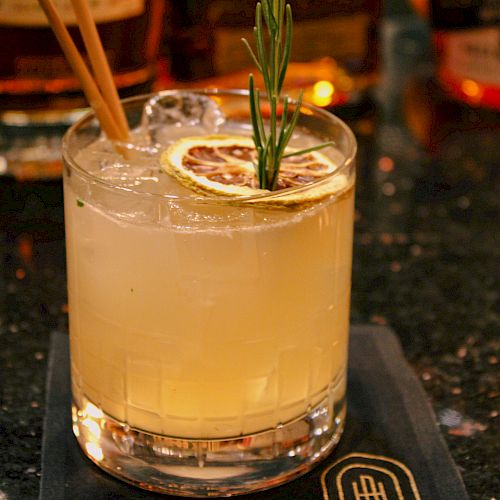 A cocktail garnished with a dried lemon slice, a rosemary sprig, and two wooden stir sticks, placed on a dark napkin on a reflective surface.