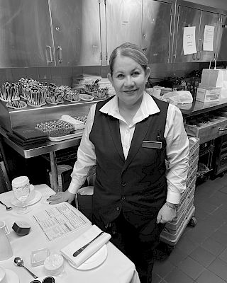 A person in a uniform stands beside a table set with dishes and utensils in a kitchen or food service area.