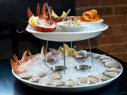 A two-tier seafood platter with oysters, shrimp, lobster, lemon slices on ice, and bowls of sides on the upper level atop a table near a brick wall.