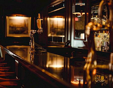 The image shows a dimly lit bar with a polished wooden counter, bar stools, beer taps, and shelves of liquor bottles in the background.