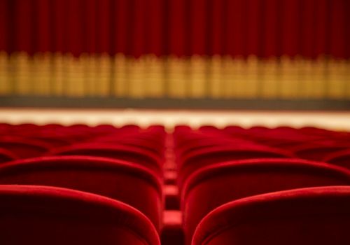 Rows of empty red theater seats facing a stage with closed red curtains in the background.