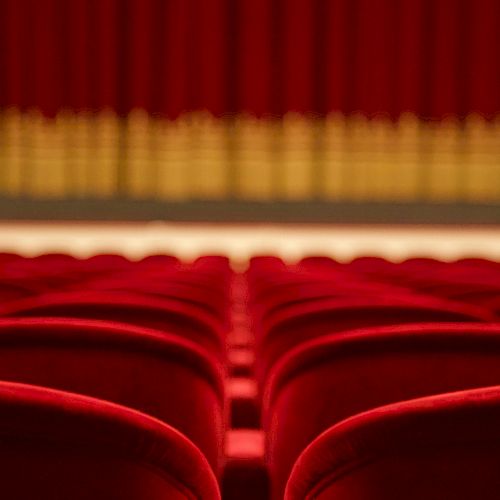 Rows of empty red theater seats facing a stage with closed red curtains in the background.